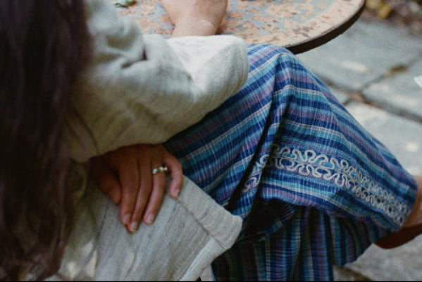 Person sitting at a table with colorful small decorations, wearing Nüwa Mano blue striped hand embroidered pants, and a silk hanten jacket