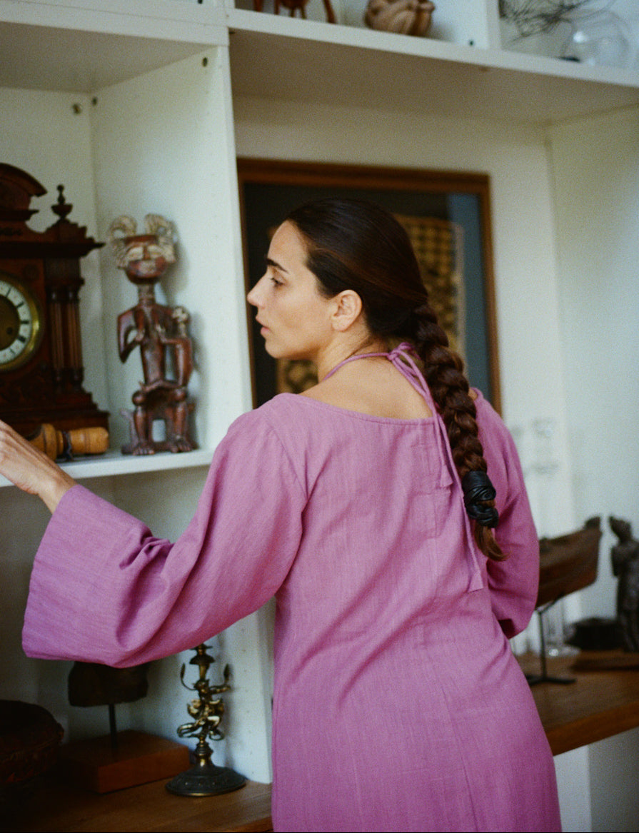 Woman in a pink dress standing in front of a shelf with various decorative items.