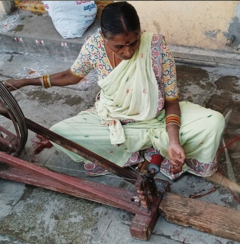 Woman spinning yarn using a traditional spinning wheel on a concrete floor.
